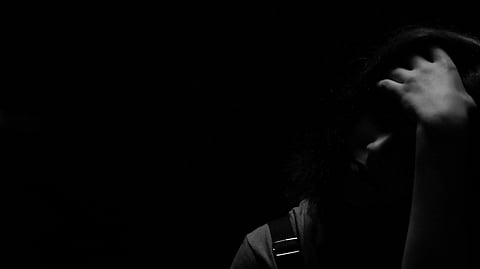 A person sits in shadowy darkness, their hand resting on their head, conveying a sense of contemplation or sadness. Dimly lit, the mood is somber.