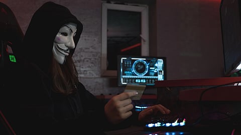 A figure wearing a hacker mask and hoodie holds a credit card while using multiple computer screens in a dim, high-tech room, conveying secrecy.