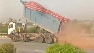 A truck is tilted on a highway, dumping a load of red dirt. Dust clouds arise as vegetation lines the road. The scene conveys motion and activity.