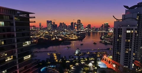 Sunset view of a vibrant city skyline with illuminated skyscrapers, a serene harbor, and a Ferris wheel, framed by nearby high-rise buildings.