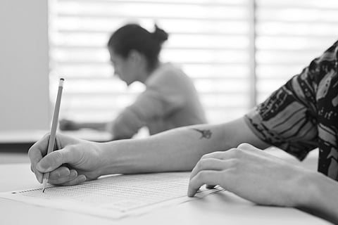 A student in a yellow patterned shirt writes with a pencil on a test paper, focusing intently. Another student is blurred in the background, creating a studious atmosphere.