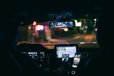 Interior nighttime view of a car dashboard. A person interacts with a smartphone and touchscreen for navigation. City lights reflect in the rearview mirror, creating a tech-savvy, futuristic ambiance.