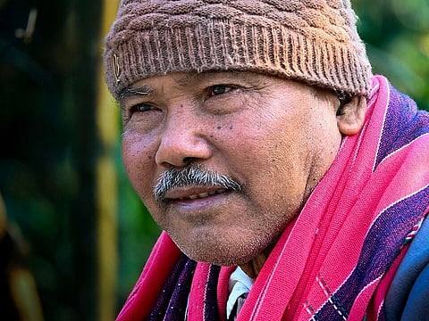 Image of India's forest man Jadav Peyang. in the image he is seen wearing a red black shawl and brown winter cap.