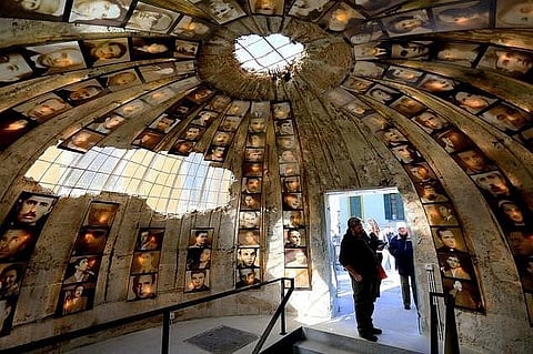 Interior of a dome-like room adorned with numerous sepia-toned portraits. A skylight at the top casts light inside, where people stand, evoking reflection.
