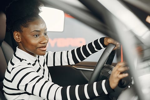 Young woman in a black and white striped top smiles while sitting in a car's driver's seat. The interior suggests a modern vehicle.