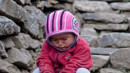 A young child in a pink-striped hat and red sweater sits pensively on stone steps, surrounded by rugged stone walls, conveying a serene, contemplative mood.