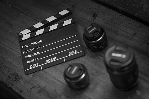 A black-and-white photo of a clapperboard titled "HOLLYWOOD" on a wooden table, surrounded by three camera lenses, suggesting a film setting.
