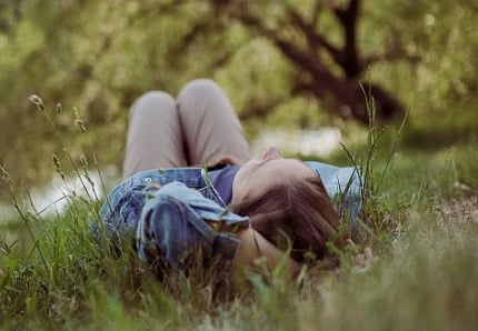 Person lying in lush grass, wearing a denim jacket, with arms behind head. Trees blurred in background, conveying relaxation and tranquility.