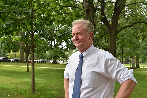 In the image Chris Van Hollen in a white shirt and blue tie stands confidently with hands on his hips in a lush park. The backdrop features green trees and grass, evoking a serene, contemplative mood.