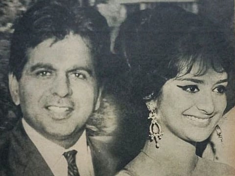 An old black and white image of Dilip Kumar and Saira Banu. the man is wearing a suit where as the woman is is wearing a saree and heavy eyeliner giving retro vibe. 