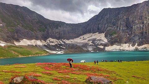 A tranquil lake surrounded by rugged mountains under a cloudy sky. A person with a red umbrella stands in a colorful meadow, evoking serenity and adventure.