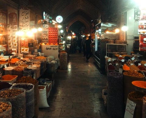 Dimly lit spice market with bright overhead lights illuminating baskets of colorful spices and herbs. Narrow aisle leads to darkened archway.