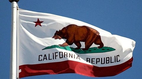 Flag of California waiving against a clear blue sky, featuring a brown grizzly bear walking on grass, with a red star above and "California Republic" text below.