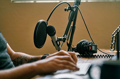 Image of a man sitting in a studio room. There is a table on which recording equipment such as mic, headphones are placed. 