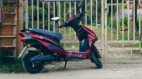 Red scooter parked in front of a gated entrance with blue signs in Bengali. Palm trees and a building are visible in the background. Peaceful setting.
