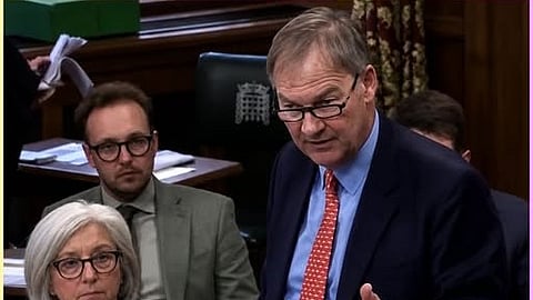 British MP Rupert Lowe presents his Inquiry report in the House of Commons wearing a blue suit and shirt with red tie as other MPs look on.