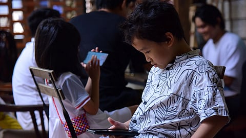 A young boy in a patterned shirt intently looks at his phone. A girl nearby is also using a device. Others in the background are blurred, creating a cozy indoor setting.