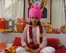 Image of tejaswi Kumari Jodha wearing a bright pink turban and floral garlands sits cross-legged on a cushion, surrounded by food and flowers, with a portrait in the background.