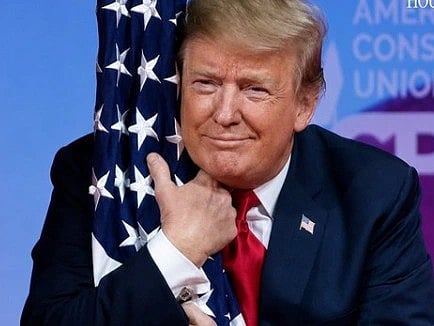 Image of Donald trump hugging the American Flag with both his hands. he is smiling in the image and is wearing his classic blue suit. 