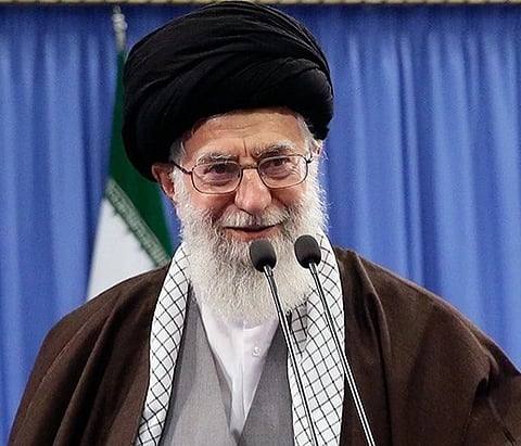 Image of  Ayatollah Ali Khamenei. in the image he is speaking in front of a mic and is smiling. 