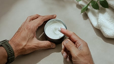 A man is taking out some beauty cream from its container. the image is minimal with a towel and a plant visible at one corner of the image. 