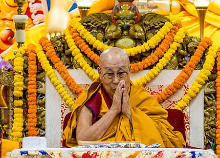 Image of  Dalai Lala with his hands folded and seated in a chair decorated with flowers. 