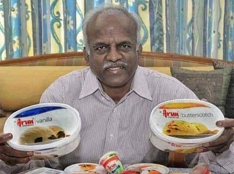 Image of R.G. Chandramogan sitting in a sofa with his most famous brand Arun ice cream containers/. various products from his ice cream business is placed in front of him. 