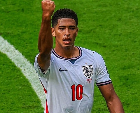 Image of a man wearing the England jersey. his one hand is raised and fists closed. he is standing on a gorund. 