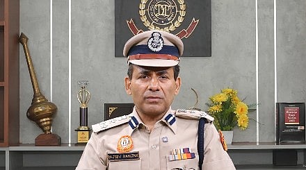Image of a senior police officer sitting his cabin and speaking in front of the camera. Behind him the delhi police logo and trophies are placed. The man is wearing his official uniform,