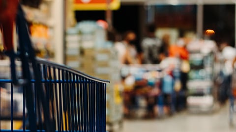 Close-up of an empty blue shopping cart in a grocery store aisle. Shelves are visible to the left, with blurry shoppers in the background.