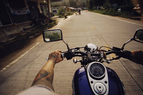 Motorcyclist's view on a sunny road, showing tattooed arms gripping handlebars. Dashboard and side mirrors visible, surroundings are blurred with motion.