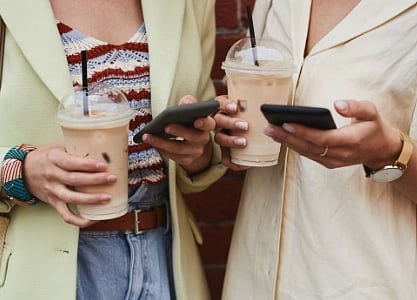 Two stylish women with iced coffee and smartphones stand against a brick wall.
