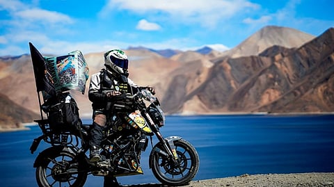 A motorcyclist in protective gear and helmet rides a bike loaded with gear, overlooking a serene blue lake and rugged mountains under a bright blue sky.