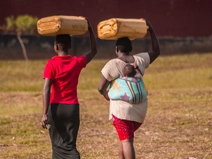 Two women in Uganda carry goods on their heads with a baby in a rural setting.

