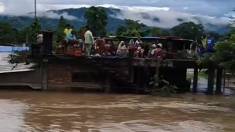In the image Assam flood is shown with people standing on the top of the house