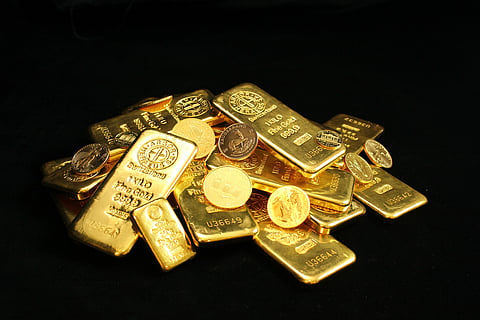 A pile of gleaming gold bars and coins stacked on a dark background, conveying wealth and luxury. The surface of the bars is engraved with text and logos.