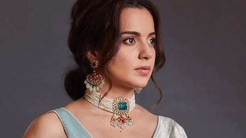 Image of Kangana ranaut in a blue saree and neck piece with an green emerald on it. In the image she is looking sideways aways from the camera. 
