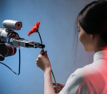 A robotic arm delicately holds a red flower as a woman interacts with it.
