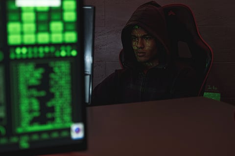 A person in a dark hoodie is intensely focused on computer screens displaying green code. The dimly lit room conveys a mysterious, tense atmosphere.