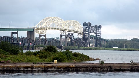 The Sault Ste Marie International Bridge spans over a calm body of water, reflecting the surrounding landscape in its surface.