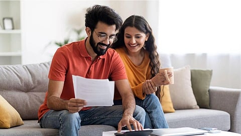 A smiling couple sits on a sofa reviewing documents. The man wears a red shirt and glasses, the woman in yellow holds a coffee mug. The mood is cheerful and relaxed.
