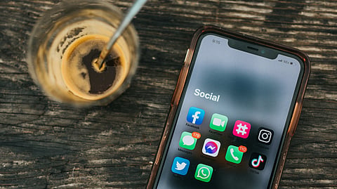 The image shows a smartphone rested on the table, with social media applications such as facebook and messenger visible on the screen. Beside the smartphone sits a glass of coffee.