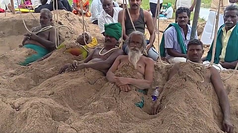 Several individuals seated on the beach, with their bodies buried in the sand, depicting a protest against water dispute
