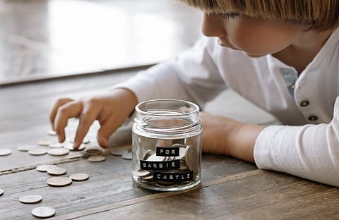 A young child collects coins in a jar labeled 'For Barbie Castle', symbolizing saving and dreams.