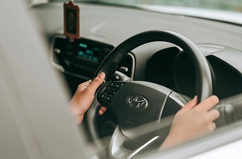 Hands grip a Toyota steering wheel inside a car, viewed through the window; dashboard controls and a hanging ornament create a focused driving mood.
