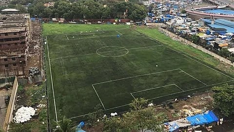 Aerial view of a green soccer field beside dense city buildings and informal settlements, with a high-rise tower under an overcast sky.