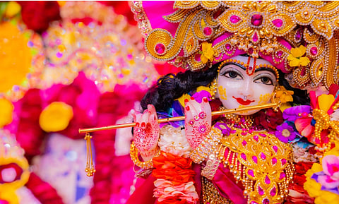Colorful close-up of Lord Krishna idol playing a flute, wearing an ornate jeweled crown and pink-gold garments, surrounded by flowers.