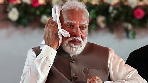 PM Modi  in a brown vest wipes his brow with a white cloth, holding glasses, with a blurred floral background. He appears contemplative and slightly weary.