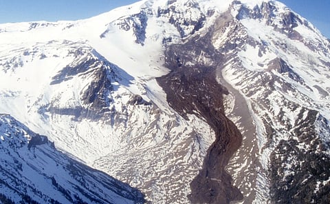 Snow-covered mountain peaks surround a dark, winding glacier or rock valley, creating a dramatic, remote alpine landscape beneath a blue sky.