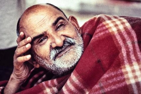Image of Neem karoli baba lying down with a blanket on him. His right hand is placed on his head to support. the picture is old